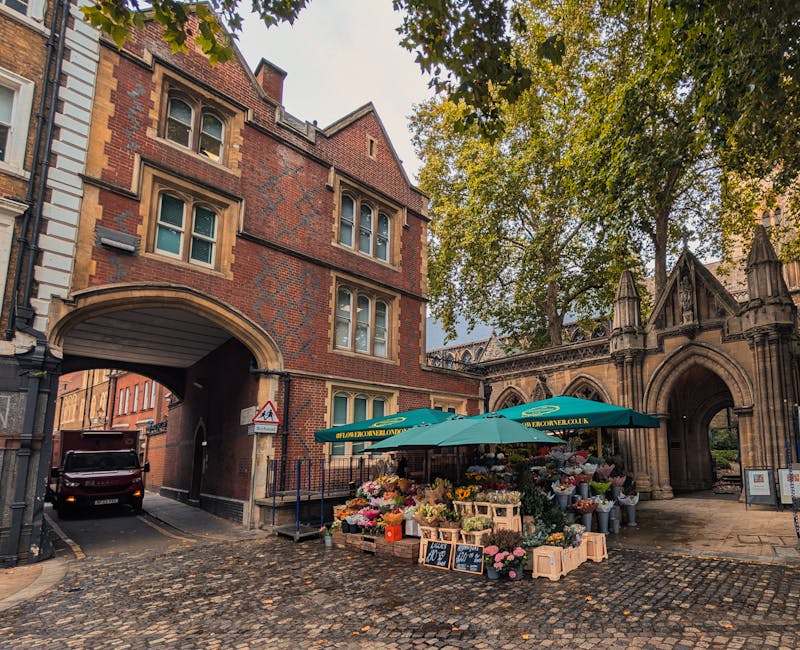 A bustling outdoor flower market situated on a cobblestone street near a historic stone archway and Gothic-style buildings. The floral stall is shaded by a teal umbrella bearing the URL flowersnorthol