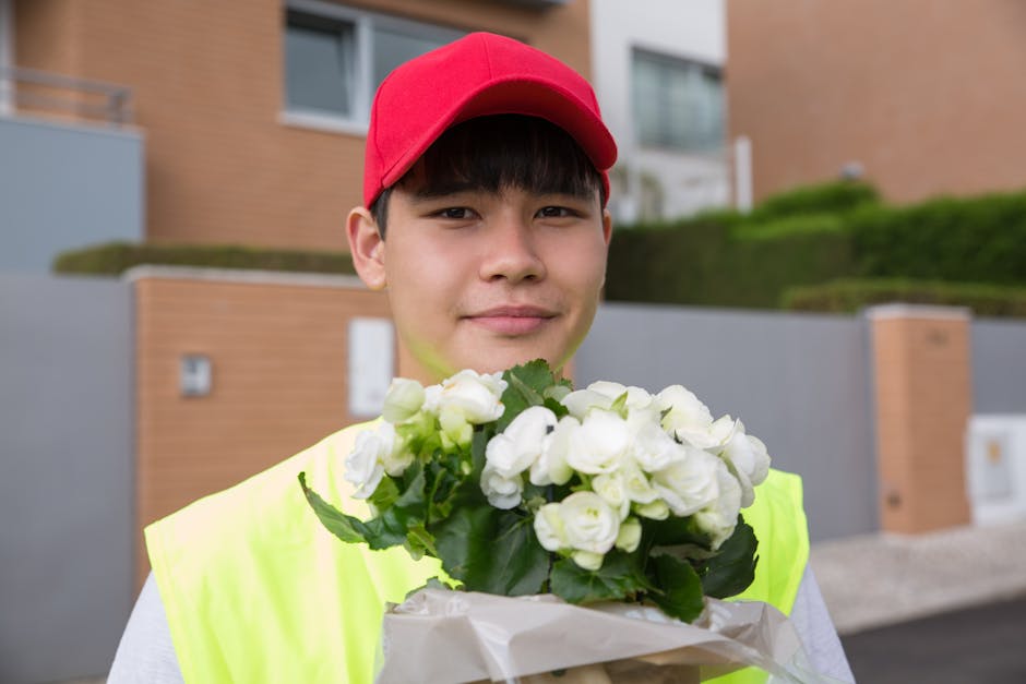 A young male flower delivery courier with light skin, wearing a red cap and a high-visibility yellow vest, stands outdoors in front of modern residential buildings with brick and grey exterior walls,