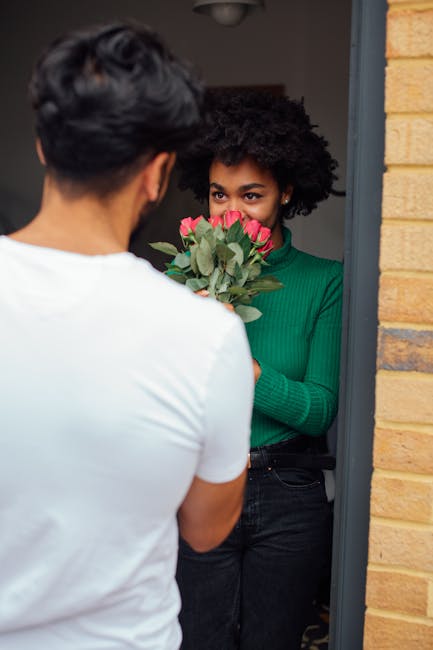 A woman with dark, curly hair wearing a green sweater holds a vibrant bouquet of pink roses and green foliage while standing in a doorway indoors. A man with dark hair, seen from behind, is handing he