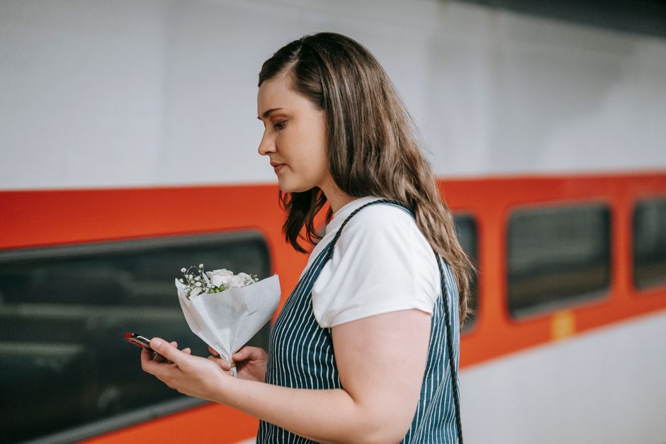 A young woman with long brown hair, wearing a white t-shirt and a blue and white striped apron, is standing on a train platform. She is holding a bouquet of fresh white flowers, wrapped in white paper