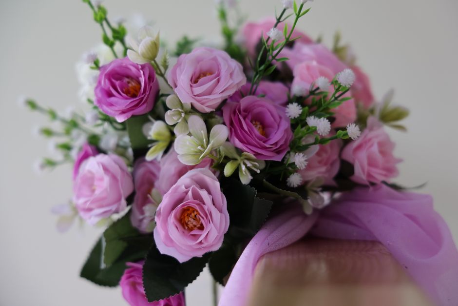 A close-up of a floral bouquet arranged with pink roses, white and light yellow small filler flowers, and green foliage, wrapped in soft pink tissue paper tied with a pink ribbon. The bouquet showcase