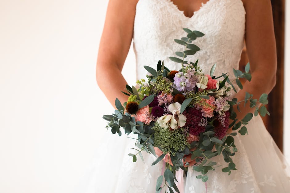 A bride in a white lace wedding gown is holding a large, lush floral bouquet featuring a mix of fresh flowers and greenery. The bouquet includes white orchids, pink and purple hydrangeas, deep burgund