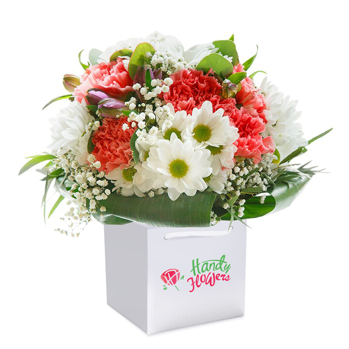 Carefully designed by a Northolt florist with a keen eye for both colour and composition, this lush bouquet stands out, upright in a sharp, clean white gift bag ready for delivery around Northolt's bustling neighbourhoods. Soft coral-pink carnations dominate, their layers of ruffled petals imparting a luxurious, tactile depth, complemented by vibrant red blooms that pulse with warmth. Next to them, fresh white daisies with rich yellow-green centers bring a gentle brightness, reminiscent of weekend mornings spent by Northala Fields. Tiny sprigs of baby's breath encircle the main flowers, their delicate, airy clusters weaving light and space into the arrangement. Look closely, and you'll spot a few purple-tinged buds, perhaps alstroemeria not yet open, peeking from the left, adding dimension and narrative to the bouquet. Deep green, broad leaves create a natural, structured base, enveloping the flowers and accentuating their vivid tones. The presentation is heightened by the elegantly sturdy ribbon handles of the gift bag, sitting against a flawless white backdrop that draws out every nuance of colour and form. This expert arrangement embodies freshness and heartfelt sentiment, perfect for local festivities, from birthdays along Ruislip Road to anniversaries celebrated in Northolt's close-knit communities-a true reflection of local floral artistry and care.