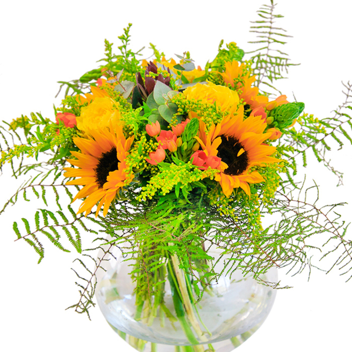 Showered in golden light, this elaborate bouquet radiates the cheerful essence of a Northolt summer, perfect for sharing joy in local homes or festive gatherings. Majestic sunflowers dominate the ensemble, their dark chocolate centers and radiant yellow-orange petals reminiscent of sunbeams streaming through the borough's parks. Surrounding them, velvety yellow garden roses bloom in delicate, tightly-packed layers, offering a sense of sophisticated beauty that echoes the classic charm of Northolt's leafy avenues. Among the blooms, clusters of shiny red hypericum berries are artfully placed, offering pops of vibrant contrast and a sense of lively festivity. The base of the bouquet brims with verdant foliage-feathery ferns, bright solidago with their clusters of tiny yellow blossoms, and slender, upright greenery, all contributing depth and texture. Soft blue-green eucalyptus leaves peek gently through the arrangement, lending a cool note amid the warmth. Nestled in a clear, fishbowl-style vase, the stems are visible, basking in the purity of fresh water, their natural lines adding to the composition's elegant architecture. Whether marking an anniversary, a birthday, or simply brightening a day in Northolt, this arrangement exudes warmth, happiness, and a genuine sense of local pride-each blossom a reflection of meticulous craftsmanship and the enduring beauty of the Northolt community.