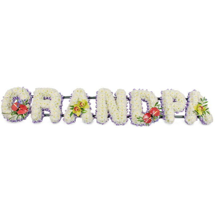 The image shows a beautifully crafted funeral flower tribute spelling "GRANDPA" in bold, three-dimensional capital letters, as though it might be placed beside a coffin or at the front of a Northolt crematorium chapel. Each letter is composed of densely packed white chrysanthemums, their small, daisy-like heads arranged so closely that they form a smooth, cushion-like surface. The cool white petals create a serene, almost luminous effect against the plain white background, symbolising purity, respect, and a peaceful goodbye. Around the base of every letter runs a neat trim of pale purple or lavender ribbon, just visible beneath the blooms. This soft edge of colour frames the word and adds a gentle, comforting contrast, as if a pastel shadow is cradling each letter. The entire word is supported by dark green bars, partly obscured but visible enough to suggest a strong framework beneath the flowers. To break the expanse of white and to personalise the design, vibrant focal sprays have been added: the 'G' and 'D' carry rich groupings of deep red anthuriums with their glossy, heart-shaped spathes and bold central spadices, arranged among lush green leaves and hints of complementary filler. The first 'A' and the final 'A' are softened with clusters of yellow-green orchids, their elegant, arching blooms offering a delicate, exotic touch. Together, these accents bring warmth, character, and individuality to the tribute. This "GRANDPA" arrangement feels like a bespoke memorial for a grandfather from the Northolt community, perhaps chosen by family meeting just off Ruislip Road, and is ideal for funeral services, wakes, or graveside tributes where a clear, heartfelt message of love and remembrance is needed.